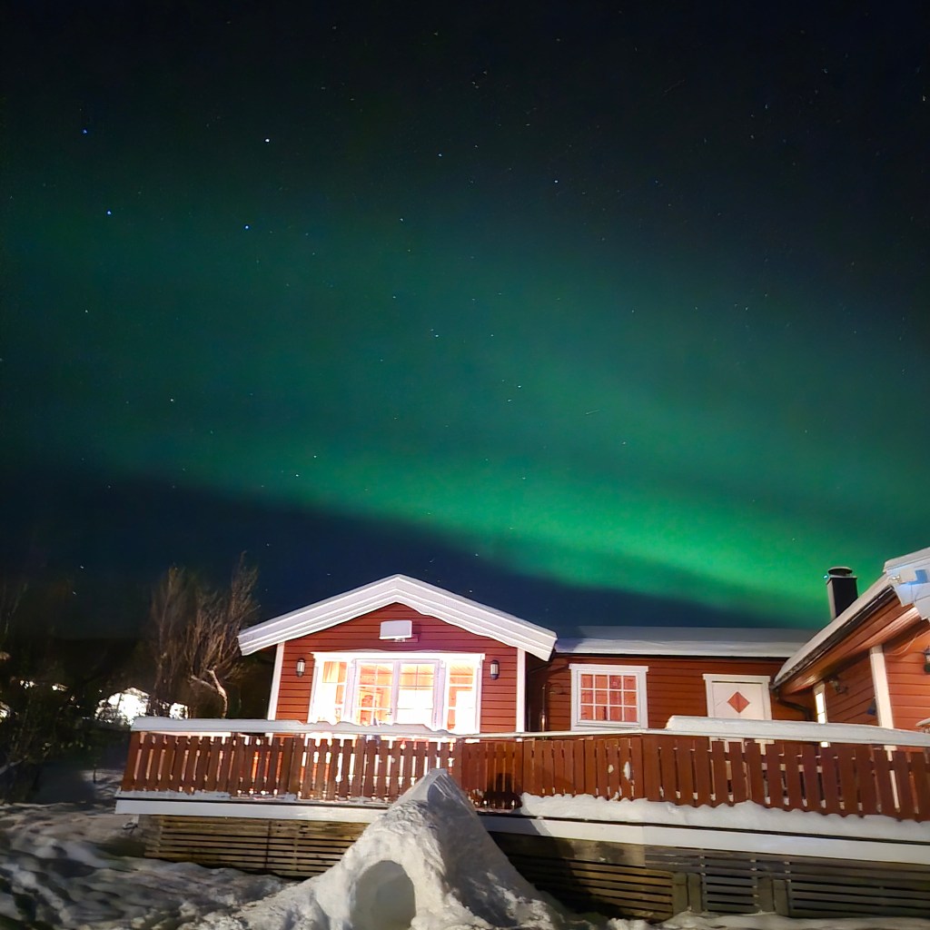 Vibrant green Northern Lights (Aurora Borealis) illuminating the night sky above a cozy red wooden cabin in a snowy winter landscape.