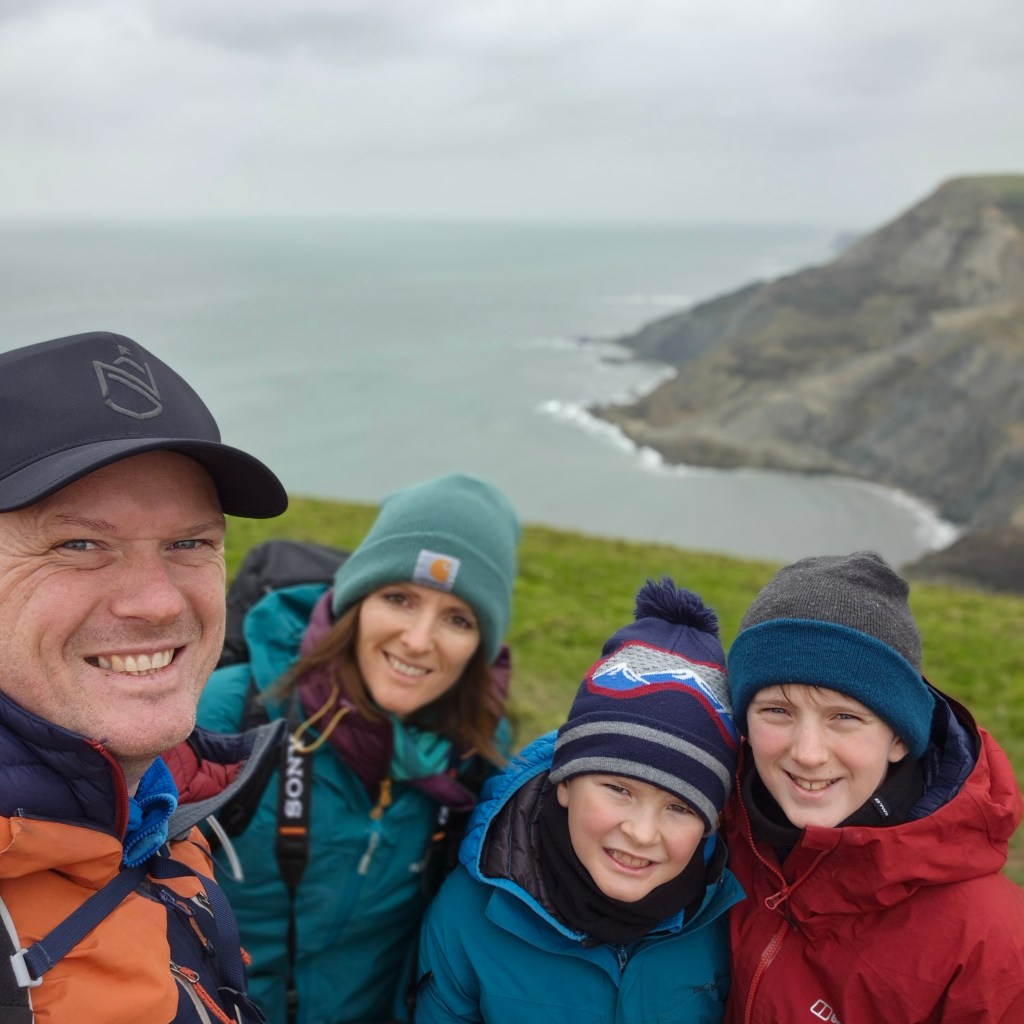 A happy family of four in colorful hiking jackets and beanies smiling for a selfie on a grassy cliff overlooking a rugged coastline.