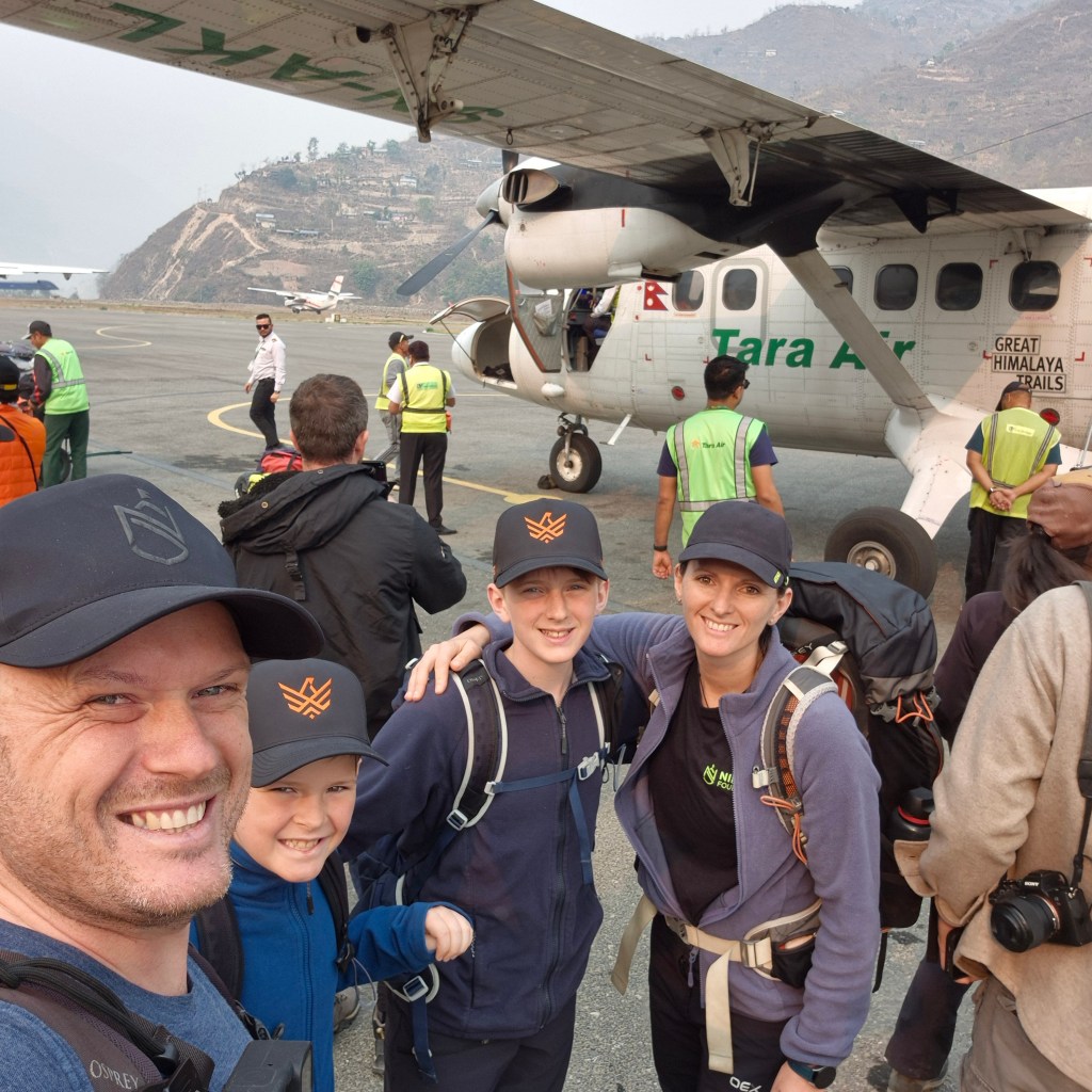 A smiling family with backpacks standing on a mountain airfield in front of a Tara Air plane during a Great Himalaya Trails trekking adventure.