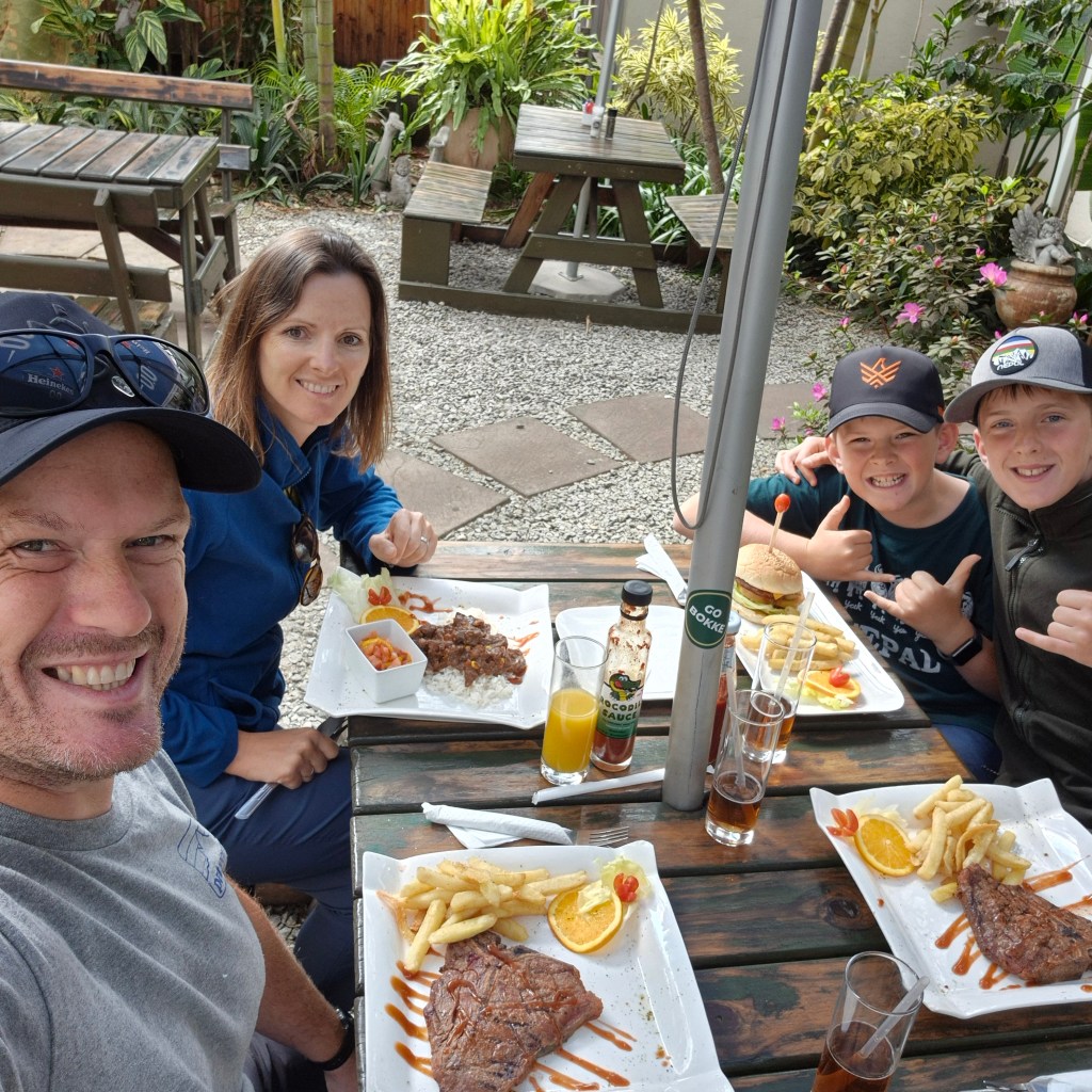 A happy family of four, including two young boys, smiles at the camera while enjoying a meal of steaks and burgers with fries at a rustic outdoor restaurant patio table surrounded by plants.