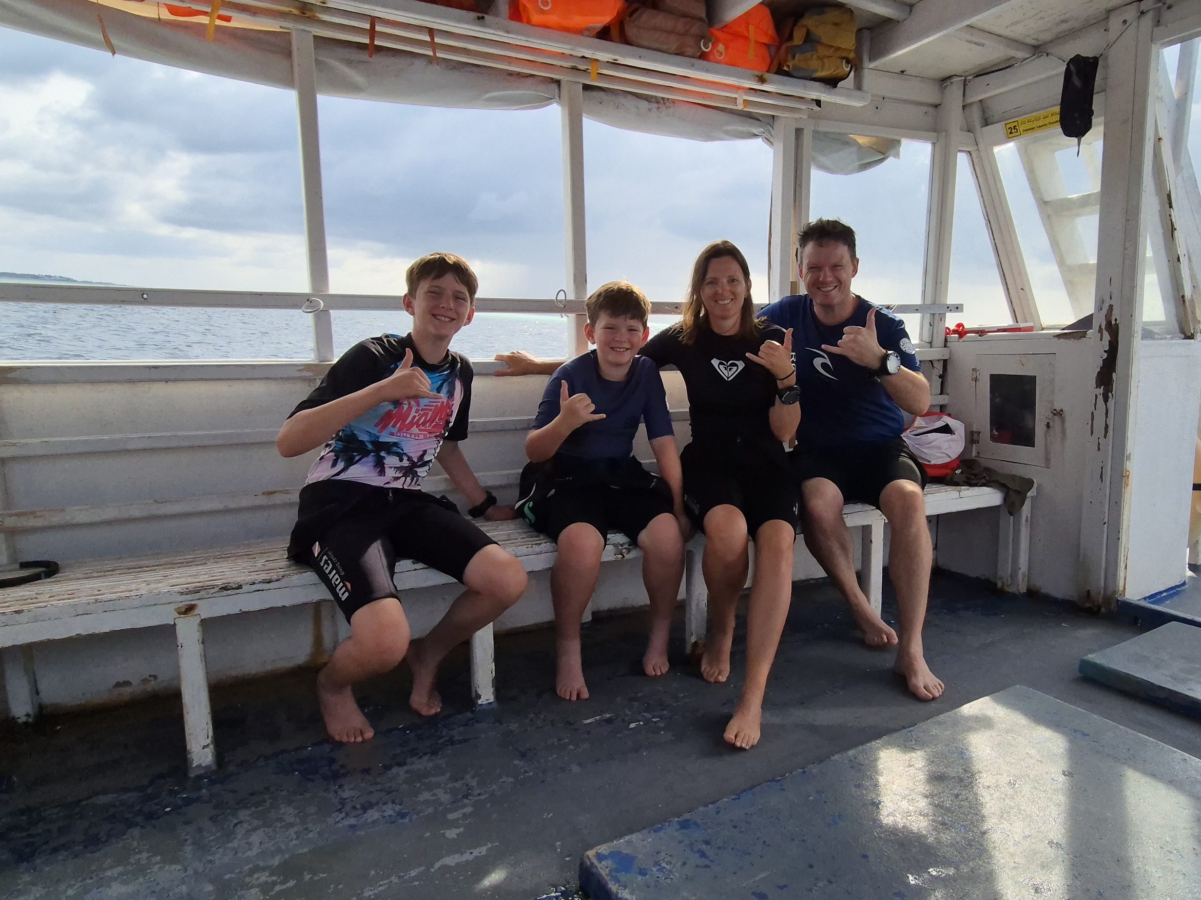 A smiling family of four wearing swim shirts and wetsuits sits barefoot on a boat bench, flashing shaka hand signs with the open ocean visible through the boat's windows.