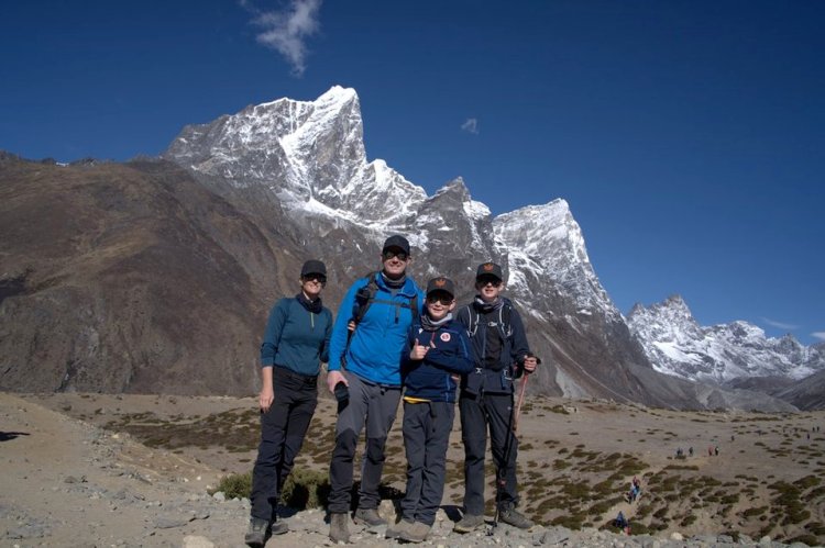 A family of four wearing hiking gear poses together on a rocky trail with towering snow-capped mountain peaks and a clear blue sky in the background during a trekking adventure.